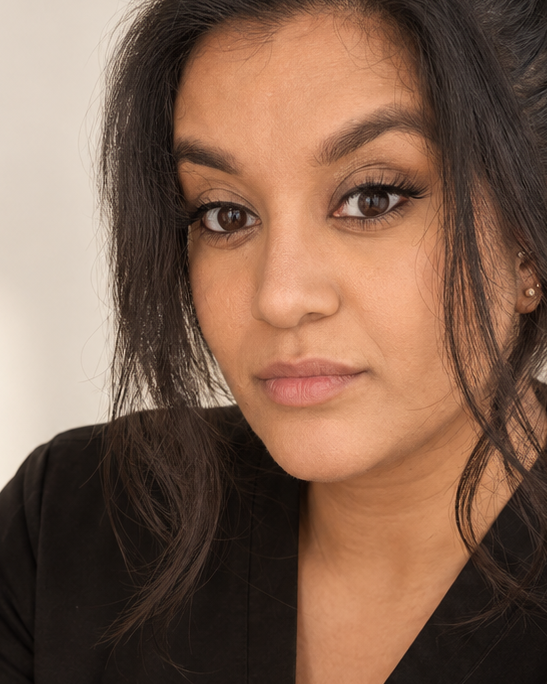 A woman with dark hair in a messy bun wearing black medical scrubs, looking directly at the camera against a warm off-white background with soft window shadows.